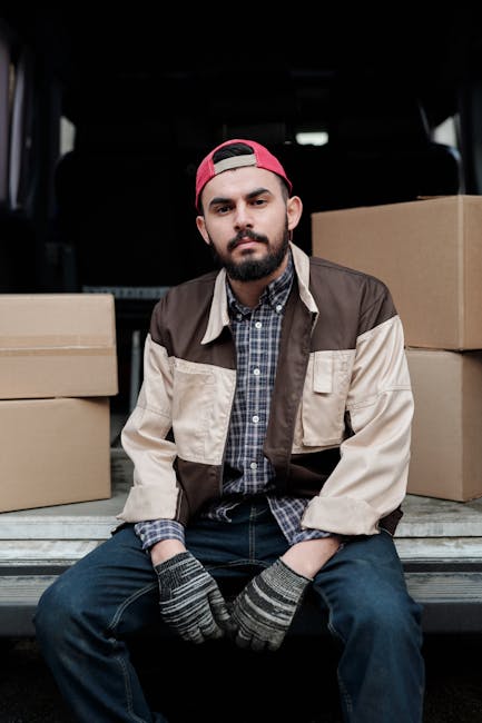 A young man with a beard, wearing a red baseball cap backwards, a brown and beige jacket over a blue checked shirt, and grey patterned gloves, sitting on the edge of a loading area at the back of a van. Behind him inside the van are large cardboard moving boxes, some stacked and others placed on the floor, indicating packing for a home relocation. The scene occurs outdoors, with the lifted tailgate of the van open, providing access to the interior, which is dark but illuminated enough to see the boxes. The man appears to be taking a break during a furniture transport and packing process, aligned with professional removals services as offered by Man with Van Wandsworth, supporting moving logistics and packing tasks involved in house removals.