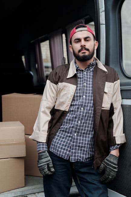 A man standing inside a moving van during a house relocation process in Wandsworth, wearing a red cap, plaid shirt, and gloves, with several cardboard boxes stacked behind him near the open rear door. The interior of the van is visible, showing additional packing materials and the vehicle's wall paneling. The scene captures the loading or unloading stage of furniture transport and packing activities involved in professional removals services by Man with Van Wandsworth, supporting efficient home relocation. Natural lighting illuminates the space, and the man's relaxed stance indicates an ongoing moving operation within a residential setting.