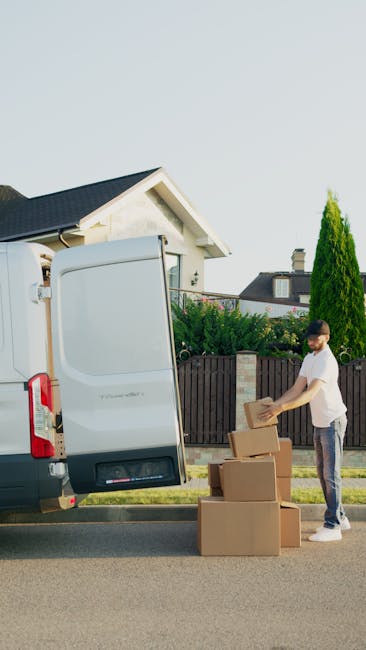 A young man with a beard, wearing a red baseball cap backwards, a brown and beige jacket over a blue checked shirt, and grey patterned gloves, sitting on the edge of a loading area at the back of a van. Behind him inside the van are large cardboard moving boxes, some stacked and others placed on the floor, indicating packing for a home relocation. The scene occurs outdoors, with the lifted tailgate of the van open, providing access to the interior, which is dark but illuminated enough to see the boxes. The man appears to be taking a break during a furniture transport and packing process, aligned with professional removals services as offered by Man with Van Wandsworth, supporting moving logistics and packing tasks involved in house removals.